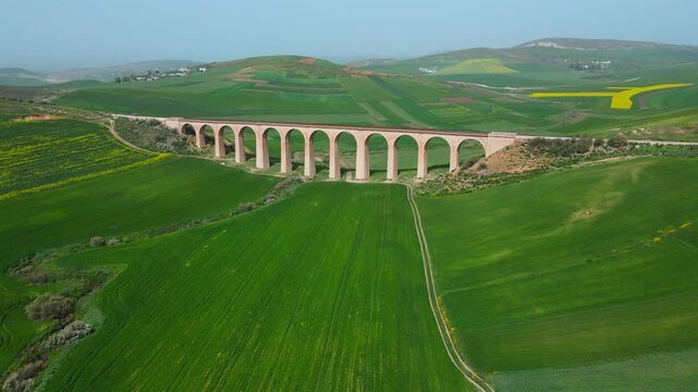 The scene is an aerial view of a bridge crossing over a valley with a train track running through it. On the other side of the valley, there are green fields and mountains in the distance.