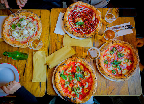 overhead shot of four pizzas with different toppings served with four beers on a rustic wooden table. Cutlery is arranged neatly for a casual dining experience.
