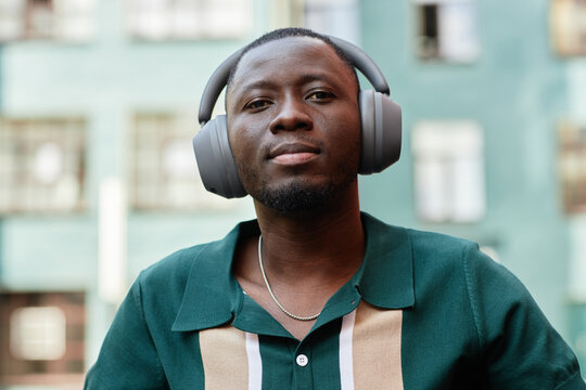 Close up portrait of Black man wearing headphones and listening to music in urban city setting looking at camera with buildings in background