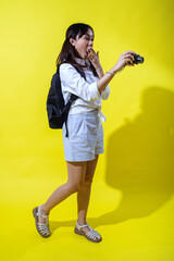 Asian woman with pigtails wearing a white shirt, and a black backpack, holding a camera and smiling enthusiastically, exuding energy and playfulness against a bright yellow background