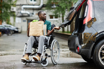 Full length scene with of young African American man using wheelchair opening car trunk in parking lot and holding groceries while doing shopping in city