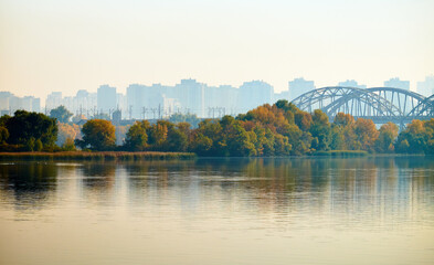 A serene riverscape with autumnal trees reflected in the water, a bridge in the distance, and a hazy city skyline