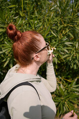 A beautiful red-haired woman in sunglasses walks through the park and sniffs the flowers on the trees on a sunny day