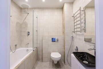 Modern, bright bathroom with beige tiles, glass shower enclosure, white bathtub, black sink, and a wall-mounted toilet. 