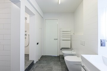 minimalist bathroom features white tiles, a sleek white door, and grey floor tiles.  A white heated towel rack and a white bidet add to the clean aesthetic.