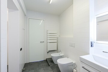 minimalist bathroom features white tiles, a sleek white door, and grey floor tiles.  A white heated towel rack and a white bidet add to the clean aesthetic. shower is visible through an open doorway. 