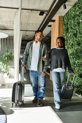 A joyful young African American couple strolls through a vibrant hotel lobby, luggage in hand.