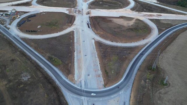 Top-down drone view of a large cloverleaf highway interchange during construction. Unfinished motorway ramps, earthworks and civil engineering infrastructure surrounded by forests and farmland before 