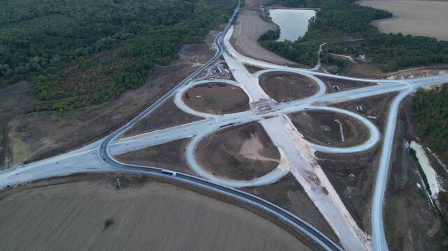 Top-down drone view of a large cloverleaf highway interchange during construction. Unfinished motorway ramps, earthworks and civil engineering infrastructure surrounded by forests and farmland before 