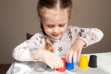 A girl in a dress at a table near a yellow wall plays sorting with colorful cups. Development of...