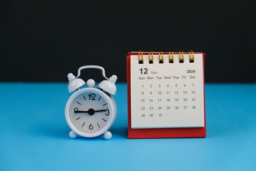 Monthly desk calendar for December 2024 year on wooden table with black alarm clock.