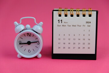 Monthly desk calendar for November 2024 year on wooden table with black alarm clock.