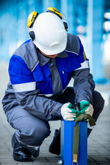 На фотографии работник в синем костюме и белой каске на голове работает руками на заводе или большом предприятии опустив голову на низ