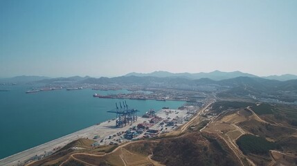 Vast Aerial View of Industrial Port Surrounded by Mountains and Blue Water Under Clear Sky, Featuring Cranes, Containers, and Coastal Landscape