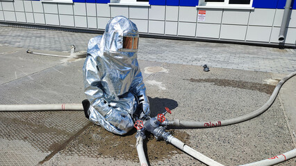 На фотографии спасатель службы мчс в специальном костюме для тушение стольного цвета идет тушить пожар в руках держит ствол с водой. Летний светлый день