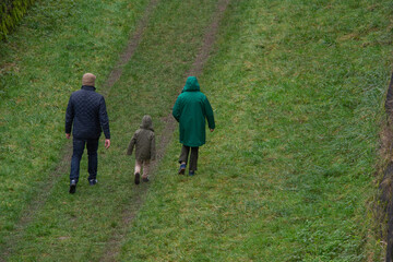Un couple vu de dos, avec un petit enfant, se promènent dans des remparts un jour de pluie . Ils sont habillés avec des imperméables verts et marche dans l'herbe 