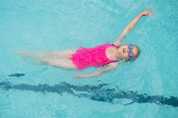 Caucasian girl in pink swimsuit and blue goggles floating on her back in a clear swimming pool