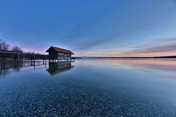 Ein Bootshaus steht einsam im absolut stillen und klaren Wasser eines See. Ein schönes Abendrot zeichnet sich am Himmel ab, und im klaren Wasser sieht man die Kiesel am Seegrund.
