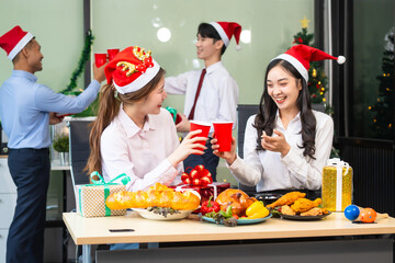 Two young businessmen and two beautiful women sit at an office desk, laughing joyfully while celebrating Christmas.They enjoy food, drinks, and exchange gifts, creating a festive and cheerful office