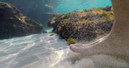 An enchanting underwater scene showcases a foot resting on the sandy ocean floor, surrounded by rocks, elegantly highlighting the submerged environment and its serene interaction with water