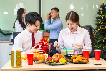 Two young businessmen and two beautiful women sit at an office desk, laughing joyfully while celebrating Christmas.They enjoy food, drinks, and exchange gifts, creating a festive and cheerful office