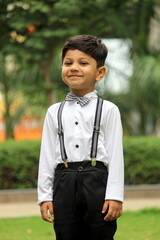 little boy is smiling and looks very cute. he is wearing formal cloths with bow tie and suspenders. He is having fun in the park in a day time. portrait of adorable child in a party wear and mood. 