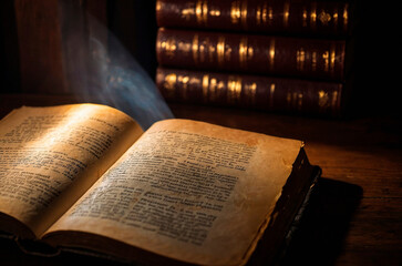 An old ancient open book laying on the table in a dim of light, blurry candles in the dark on the background. Christianity religion Holy Bible, Gospel, Testament, spiritual literature, sacred text.