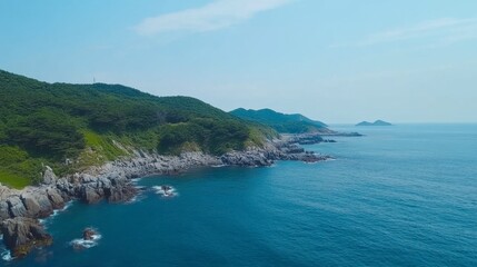 Fototapeta premium Scenic Aerial View of Rocky Coastline with Lush Green Hills and Calm Blue Ocean Under Clear Sky on a Sunny Day