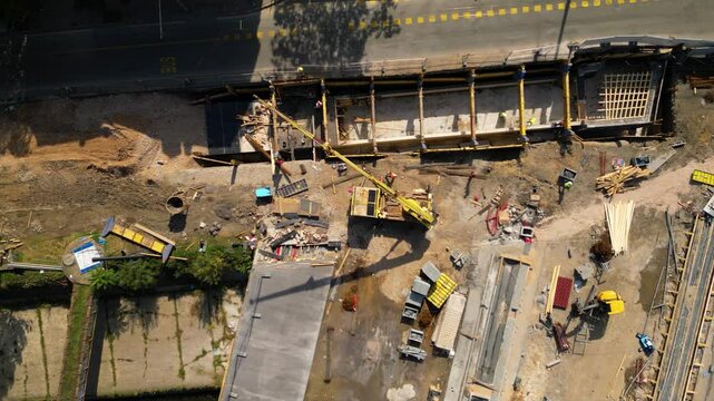 Top-down aerial view of an active urban construction site beside a roadway, with concrete works, formwork, scaffolding, a boom lift and building materials arranged around an unfinished structural sect