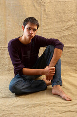 Young brunette in blue jeans and a purple sweater sits barefoot against a background of crumpled beige fabric