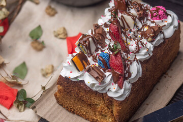   Close-up of a beautifully decorated dessert loaf cake with a rich texture, adorned with cream, fresh strawberries, and colorful chocolates. Set on a rustic table with natural elements.