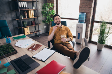 Full length photo of handsome young guy eat peanuts legs table dressed yellow shirt formalwear comfortable startup office room interior