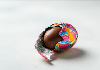 Partially unwrapped colorful foil chocolate Easter egg on white surface. Creative Easter design