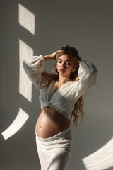 Fototapeta premium Beautiful pregnant woman in special white trousers and top. Advertisement of comfortable pregnancy Happy blond pregnant girl stand in studio with sun light. Long curly hair. Shadows on floor