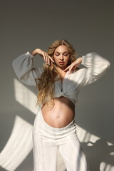 Fototapeta premium Beautiful pregnant woman in special white trousers and top. Advertisement of comfortable pregnancy Happy blond pregnant girl stand in studio with sun light. Long curly hair. Shadows on floor