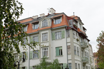 A charming European apartment building boasts a vibrant green facade adorned with decorative details and complemented by a striking red roof, all of which is beautifully surrounded by lush trees