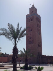 Fototapeta premium The iconic Koutoubia Mosque rises majestically against a bright blue sky, surrounded by swaying palm trees and a circular stone fountain in the heart of Marrakech.