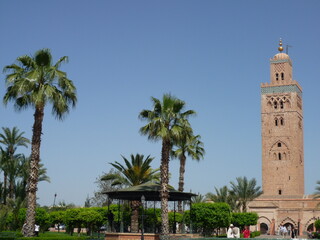 Obraz premium The iconic Koutoubia Mosque rises majestically against a bright blue sky, surrounded by swaying palm trees and a circular stone fountain in the heart of Marrakech.
