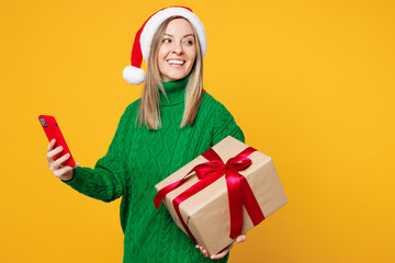 Merry young woman wear green sweater Santa hat hold present box with gift use mobile cell phone look aside isolated on plain yellow background. Happy New Year, celebration Christmas holiday concept.