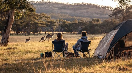 Obraz premium A couple sits on folding chairs outside their tent, enjoying a morning coffee while observing a group of kangaroos grazing in the distance.