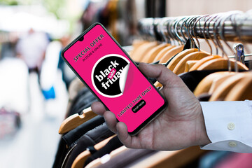 man's hand showing smartphone with black Fiday offers in front of a clothes shop