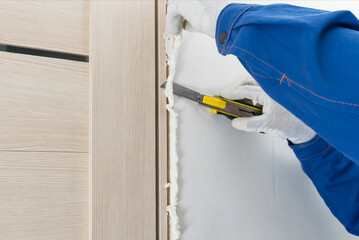 A worker in gloves cuts off excess mounting foam insulation from a door frame with a knife
