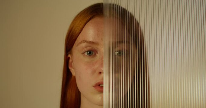 A red-haired woman with freckles looks at the camera with a serious look standing on a studio background. Half of her face is covered with plexiglass