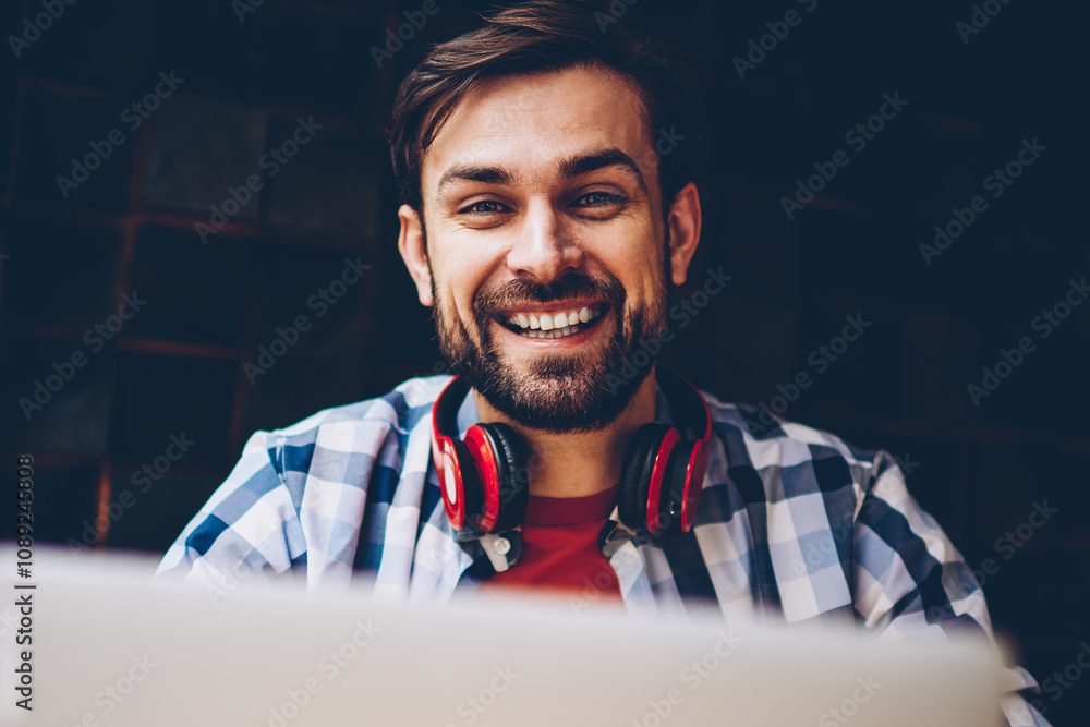 Poster portrait of at successful bearded male freelancer smiling at camera while working remotely at laptop