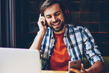 Positive bearded young man in modern headphones singing favourite audio song from playlist downloaded on smartphone.Happy hipster guy listening music from radio player on telephone sitting at laptop