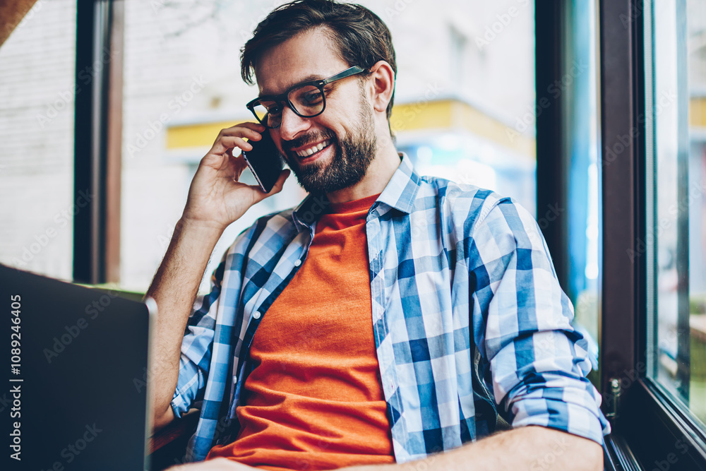 Poster happy bearded young man in eyeglasses communicating on smartphone while updating software on modern 