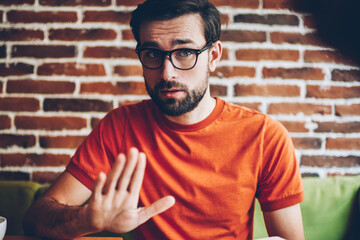 Sceptical hipster guy dressed in trendy orange t-shirt arguing during meeting.Young man with beard and eyeglasses for vision correction showing sign stop sitting in stylish coworking space