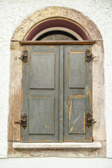 Asymmetrischer verwitterter Fensterladen aus Holz mit Eisenbeschlägen an historischem Gebäude mit Rundbogenfenster