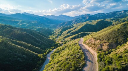 Fototapeta premium Aerial perspective of a winding mountain road nestled among lush green hills and dramatic peaks under a bright sky with scattered clouds.