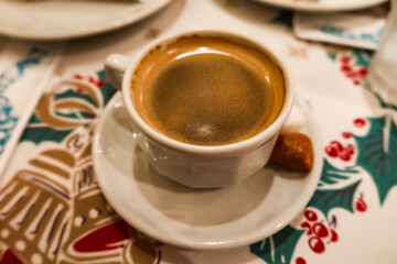 close up of hot black coffee with foam on top with a brown sugar on the side on a table cloth with Christmas red and green pattern